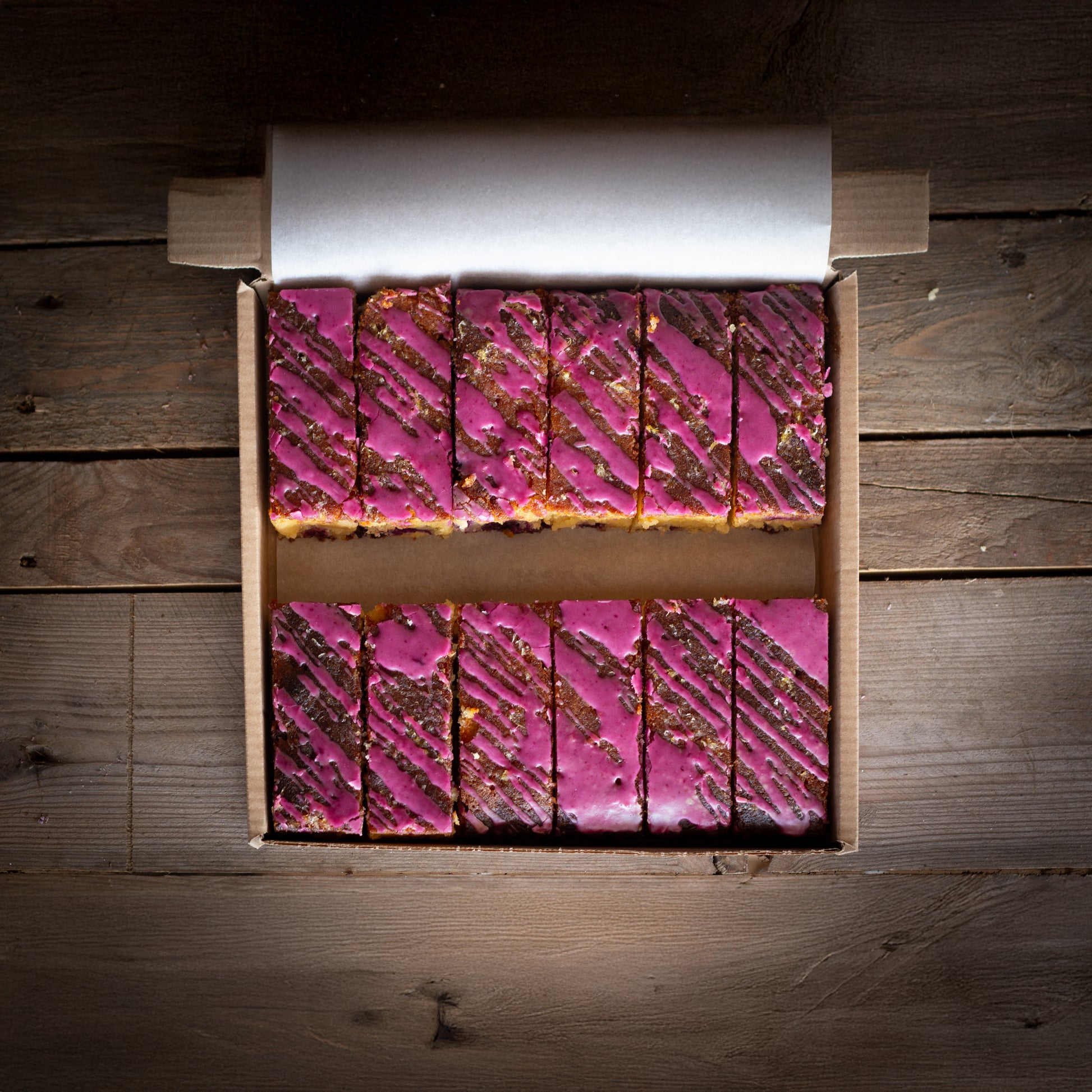 A box of sliced lemon and blueberry cake with blueberry icing on a wooden surface.